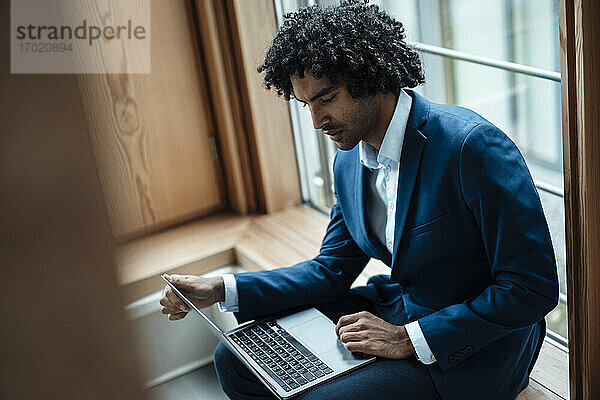 Junge männliche Fachkraft  die einen Laptop benutzt  während sie im Büro am Fenster sitzt