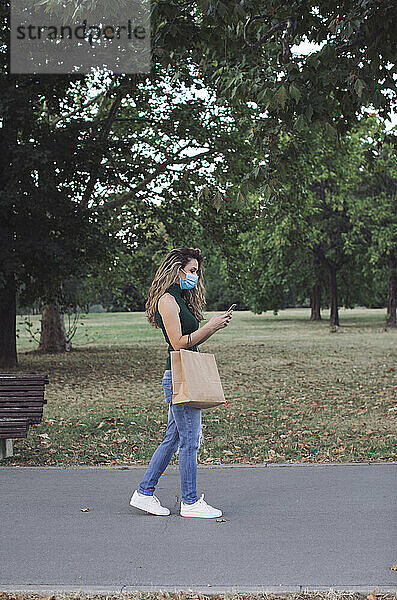 Junge Frau mit Papiertüte und Mobiltelefon beim Spaziergang in einem öffentlichen Park während COVID-19