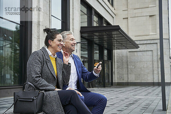 Fröhliche männliche und weibliche Unternehmer schauen weg  während sie mit ihrem Smartphone vor einem Gebäude sitzen