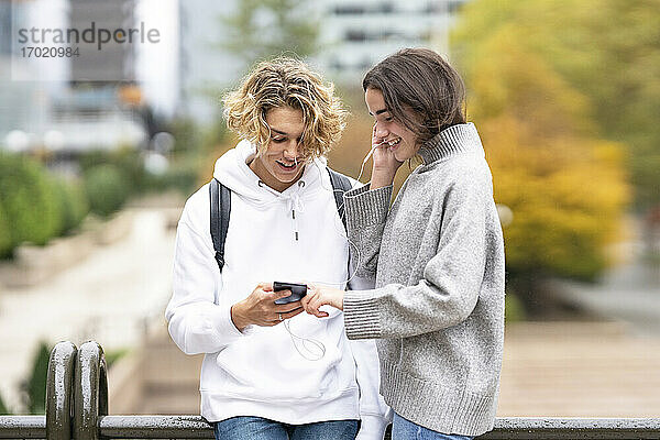 Freunde hören Musik über ihr Smartphone mit Kopfhörern  während sie sich im Park an ein Geländer lehnen