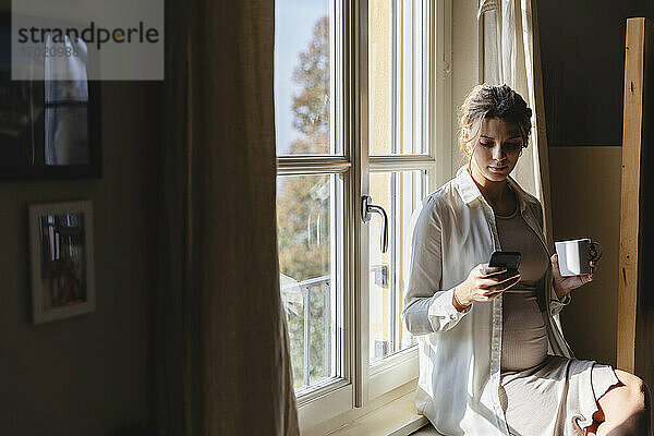 Schwangere Frau mit Kaffeetasse Textnachrichten auf Handy gegen Fenster