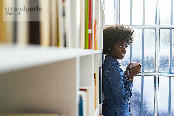 Junge Frau mit Kaffeetasse  die sich zu Hause an ein Bücherregal lehnt