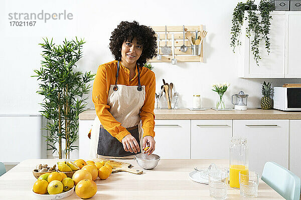 Lächelnde Frau  die eine Orange in einer Schüssel auspresst  während sie in der Küche steht