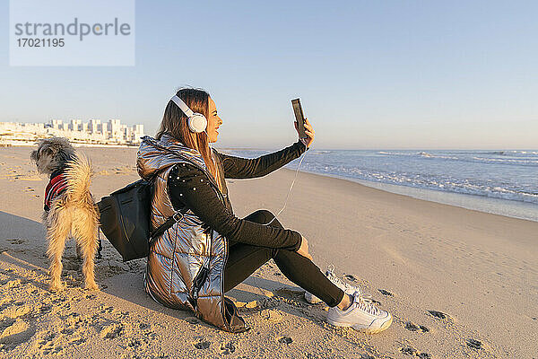 Frau  die ein Selfie macht  während sie mit ihrem Hund am Strand gegen den klaren Himmel bei Sonnenuntergang sitzt