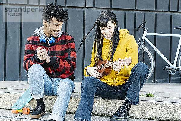 Lächelnde Frau spielt Gitarre  während sie neben einem männlichen Freund auf dem Fußweg sitzt