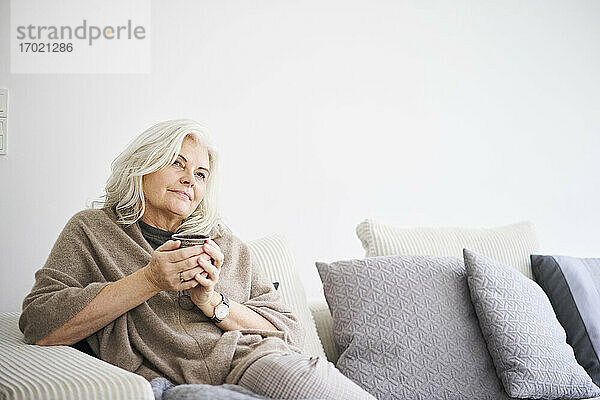Nachdenkliche Frau im Ruhestand hält eine Kaffeetasse  während sie auf dem Sofa vor einer weißen Wand in einer Wohnung sitzt