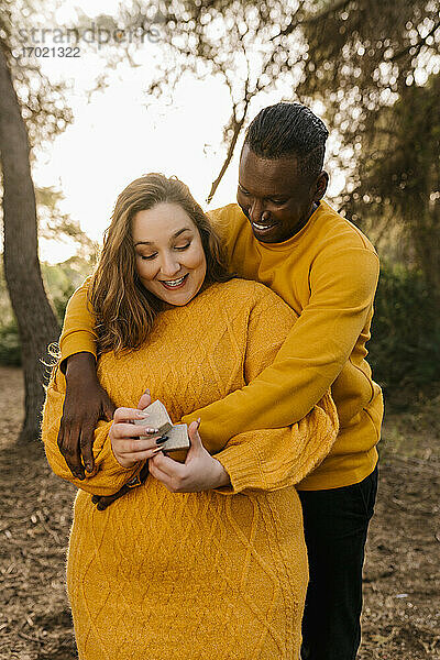 Mann umarmt Frau lächelnd und hält Verlobungsring Box im Wald