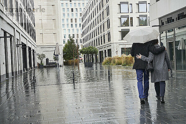 Geschäftsfrau und älterer Geschäftsmann  die sich unter einem Regenschirm in der Stadt während der Regenzeit umarmen
