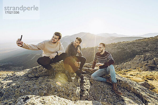 Junger Mann  der ein Selfie mit Freunden macht  während er an einem sonnigen Tag auf einem Berg sitzt