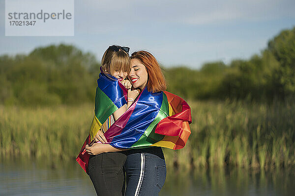 Junges lesbisches Paar mit Regenbogenfahne umarmt sich am See stehend