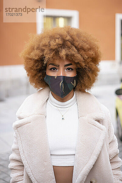 Mittlere erwachsene Frau mit Afro-Haar  die eine Gesichtsmaske trägt und an einem Gebäude steht