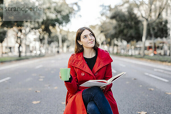 Junge Frau in roter Jacke mit Kaffeetasse träumt  während sie ein Buch auf der Straße hält