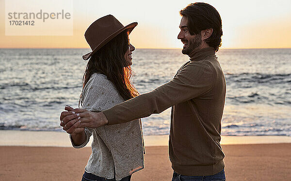 Lächelndes Paar hält sich beim Tanzen am Strand während des Sonnenaufgangs an den Händen