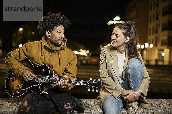 Freundin hört ihrem Freund zu  der singt und Gitarre spielt  während er auf einer Mauer sitzt