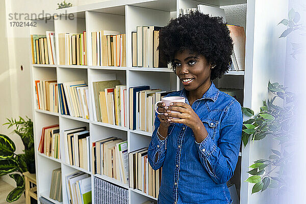 Frau schaut weg  während sie Kaffee trinkt und sich zu Hause an ein Bücherregal lehnt