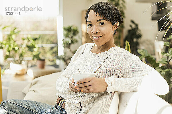Lächelnde junge Frau hält eine Kaffeetasse  während sie zu Hause auf dem Sofa sitzt