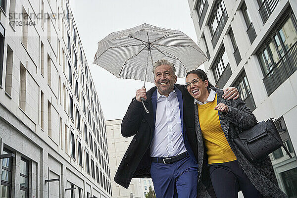 Fröhliche Geschäftsfrau und älterer Geschäftsmann mit Regenschirm beim Laufen in der Stadt während der Regenzeit