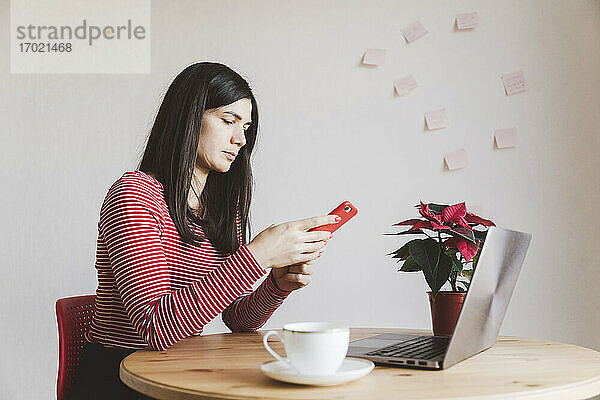 Frau mit Laptop  die ein Mobiltelefon benutzt  während sie zu Hause im Büro sitzt