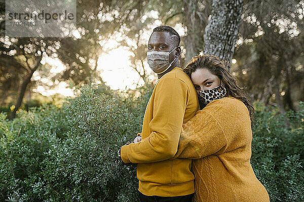 Frau mit Gesichtsmaske umarmt Mann während Covid-19 im Wald stehend
