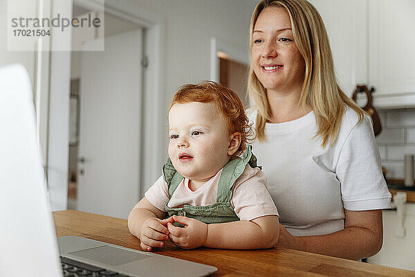 Frau mit kleiner Tochter benutzt Laptop am Küchentisch