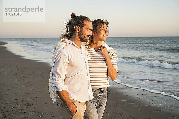 Lächelndes junges Paar schaut weg  während es im Urlaub am Strand spazieren geht