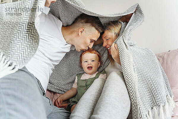 Glückliche Familie mit kleiner Tochter  die sich unter einer Decke auf dem Sofa versteckt
