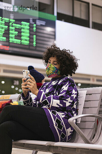 Junge Frau mit Gesichtsmaske  die ein Mobiltelefon benutzt  während sie auf einem Flughafen während des Covid-19 sitzt