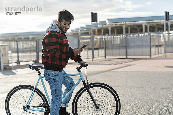 Junger Mann mit Kapuzenhemd lächelt  während er ein Mobiltelefon benutzt und mit dem Fahrrad auf der Straße steht