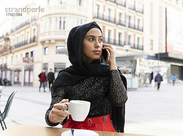 Porträt einer jungen schönen Frau mit schwarzem Hidschab  die in einem Straßencafé ein Smartphone benutzt