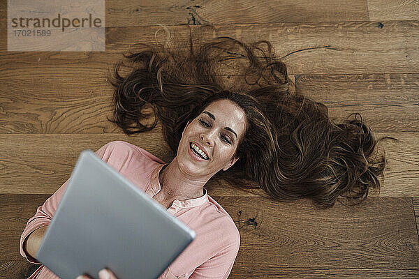Glückliche reife Frau  die ein digitales Tablet benutzt  während sie in der Wohnung auf dem Rücken liegt