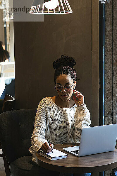 Schöne junge Frau schreibt in Tagebuch  während sie mit Laptop im Café sitzt