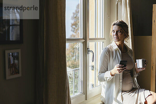 Junge schwangere Frau mit Kaffeetasse und Handy in der Hand  während sie durch das Fenster ihres Hauses schaut