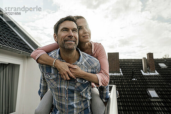 Lächelnder reifer Mann  der eine Frau huckepack auf einem Balkon gegen den bewölkten Himmel trägt