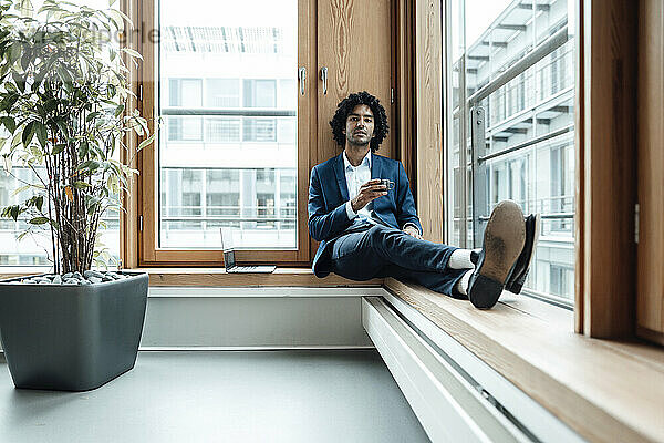 Selbstbewusster männlicher Unternehmer  der eine Kaffeetasse hält  während er im Büro am Fenster sitzt