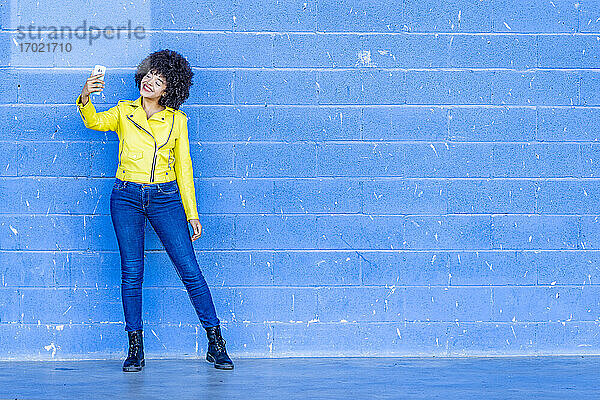 Glückliche modische Frau in gelber Lederjacke nimmt Selfie gegen blaue Wand