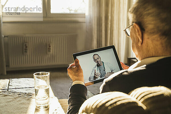 Männlicher Arzt berät einen älteren Mann per Videoanruf über ein digitales Tablet im Wohnzimmer