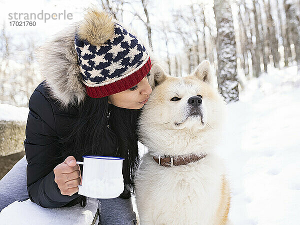 Mittlere erwachsene Frau mit Kaffeetasse küsst Hund  während sie auf einer Bank im Schnee sitzt