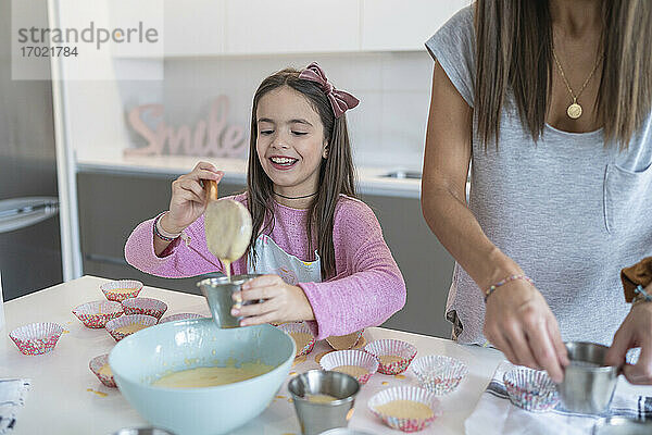 Lächelnde Tochter schöpft Teig in Cupcake-Halter von Mutter in Küche