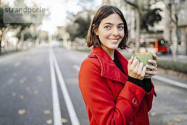 Glückliche junge Frau  die nachdenklich ist und eine Kaffeetasse auf der Straße hält
