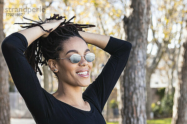 Modische junge Frau mit Sonnenbrille und Händen in Dreadlocks im Park
