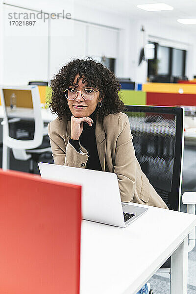 Junge Geschäftsfrau mit Hand am Kinn sitzt mit Laptop am Schreibtisch im Büro