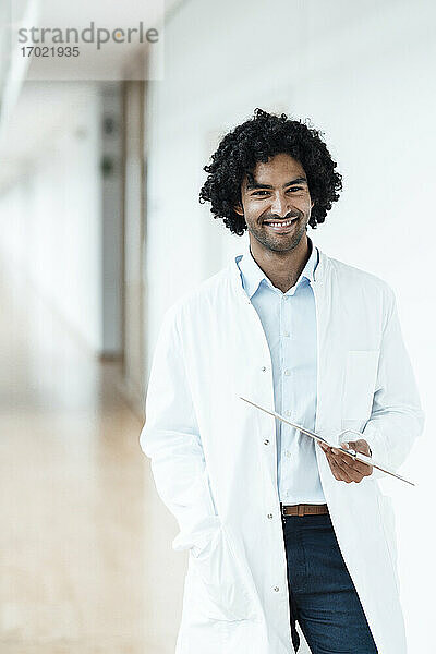 Lächelnder junger Mann im Gesundheitswesen  der ein Klemmbrett hält  während er im Krankenhausflur steht