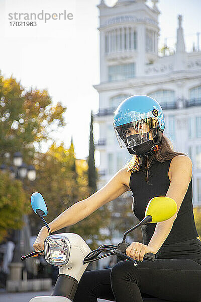 Junge Frau mit Maske und Helm beim Motorradfahren in der Stadt