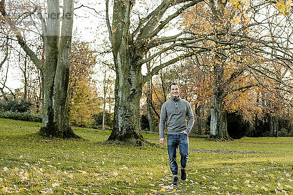 Lächelnder Mann  der im Gras vor Bäumen in einem öffentlichen Park spazieren geht