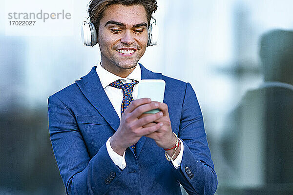 Glücklicher männlicher Berufstätiger  der ein Smartphone benutzt  während er Musik vor einem Bürogebäude hört