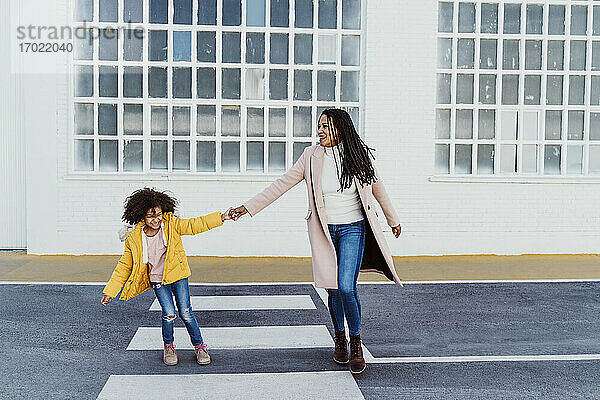 Fröhliche Mutter und Tochter halten sich an den Händen  während sie auf der Straße gegen ein Gebäude laufen
