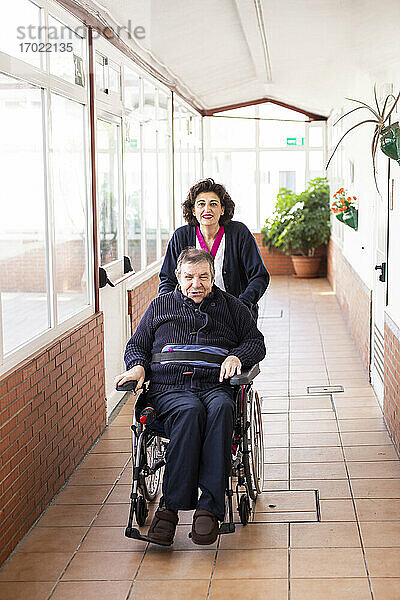 Lächelnde weibliche Betreuerin  die einem behinderten Mann im Rollstuhl sitzend im Korridor hilft