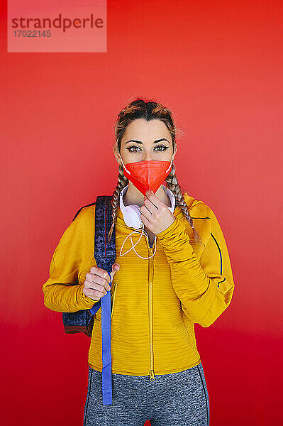 Sportlerin mit Rucksack beim Einstellen der Gesichtsmaske  während sie an einer roten Wand steht