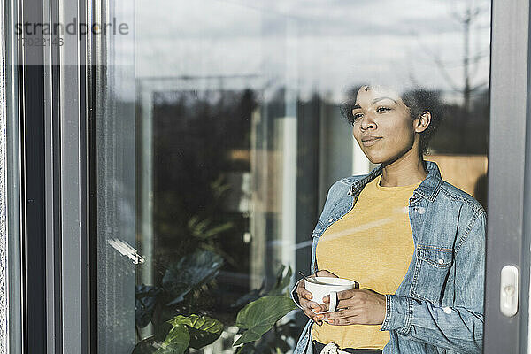 Frau schaut durch ein Fenster  hält eine Kaffeetasse