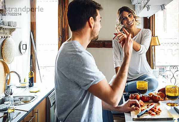 Junger Mann bereitet Essen zu und gestikuliert mit Freundin in der Küche zu Hause